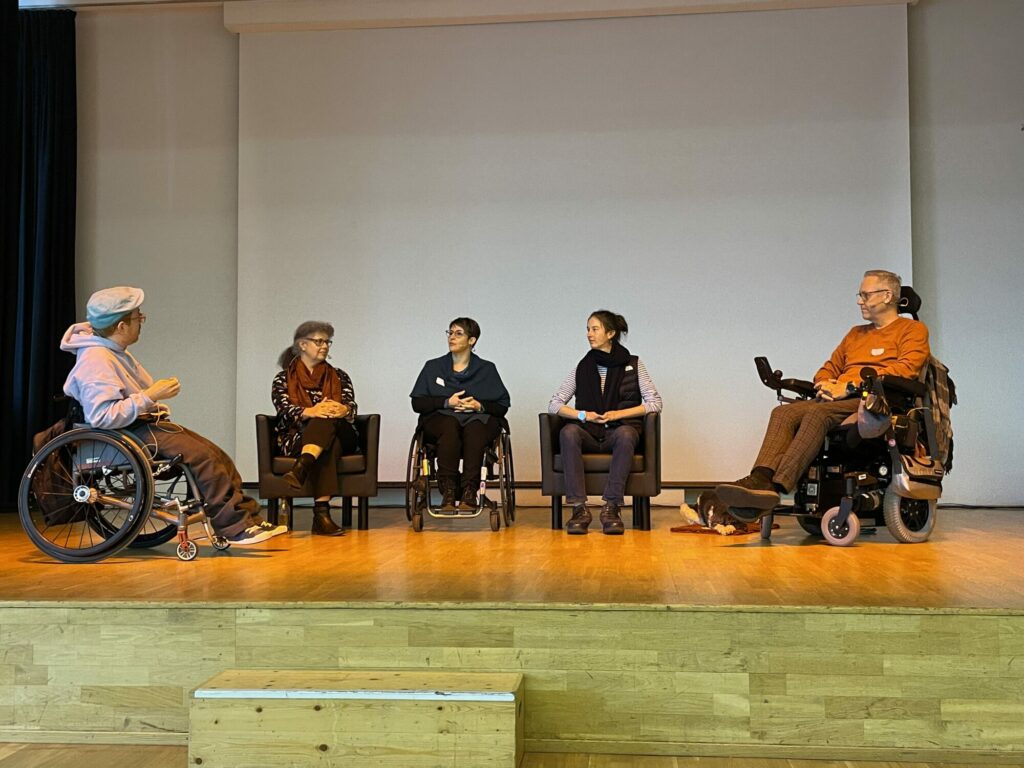 Fünf Personen sitzen auf einem Podium und diskutieren miteinander. Drei von ihnen sitzen in einem Rollstuhl