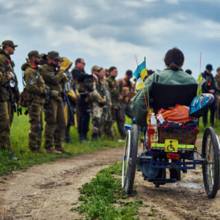 Ein junger Mann im Rollstuhl vor dem Hintergrund einer Gruppe von Soldaten in Militäruniform.