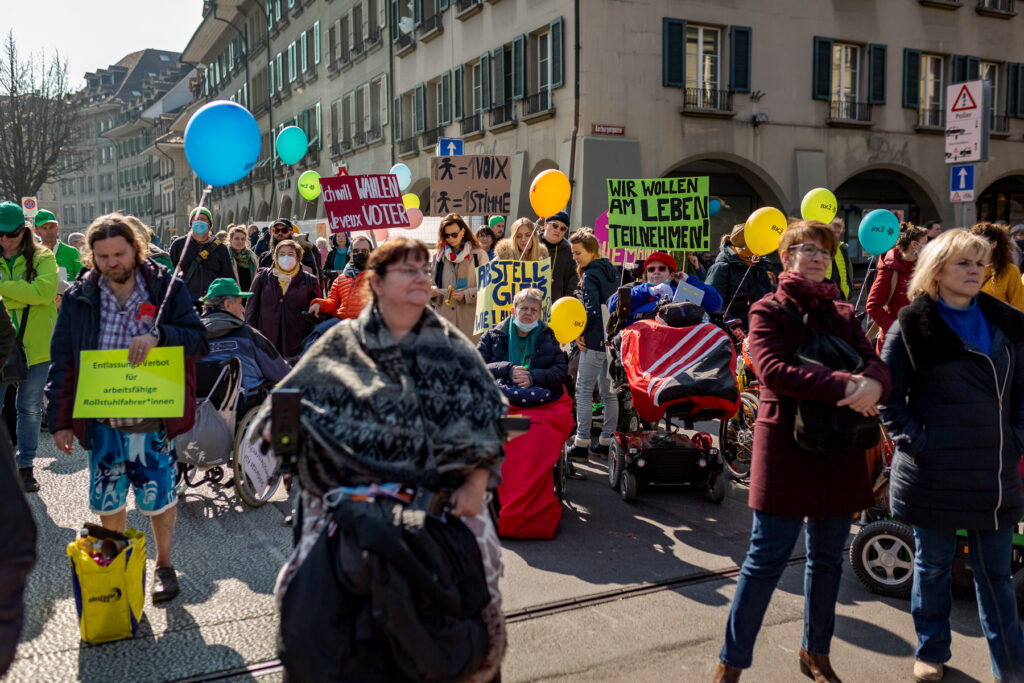 Des personnes, dont certaines en fauteuil roulant, manifestent avec des pancartes et des ballons pour défendre leurs droits.