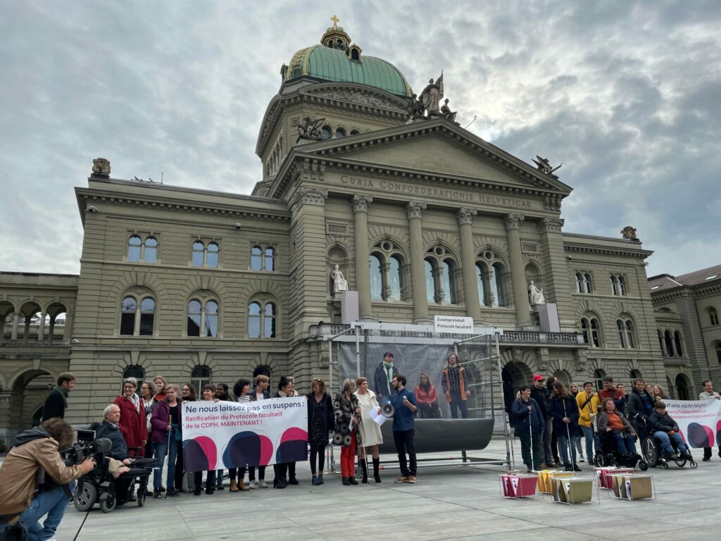 Un groupe de personnes, dont certaines en fauteuil roulant, se tiennent devant le Palais fédéral avec des banderoles.