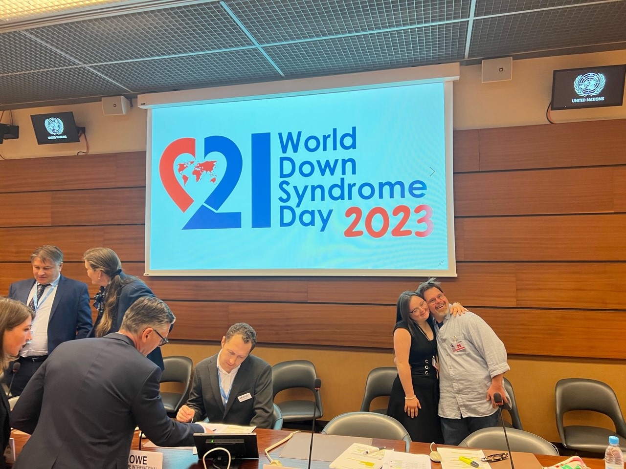 Plusieurs personnes se tiennent dans une grande salle avec pour arrière plan en projection le logo de la Journée mondiale de la trisomie 21.